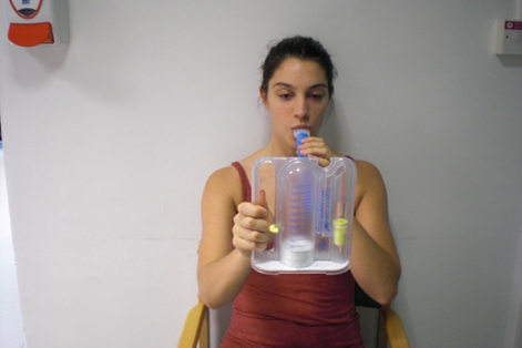 Photo of a patient seated, holding a spirometer in front of them with one hand. The spirometer is a plastic device about the size of a dinner plate with a flexible tube sticking out. The patient has the tube in their mouth.