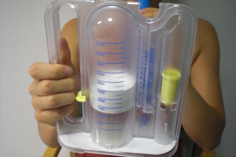 Photo showing the patient taking a breath through the device. The device has a vertical tube with divisions marked on it. Inside is a white disc that can move freely up and down the vertical tube. It's raised about an inch up the tube.
