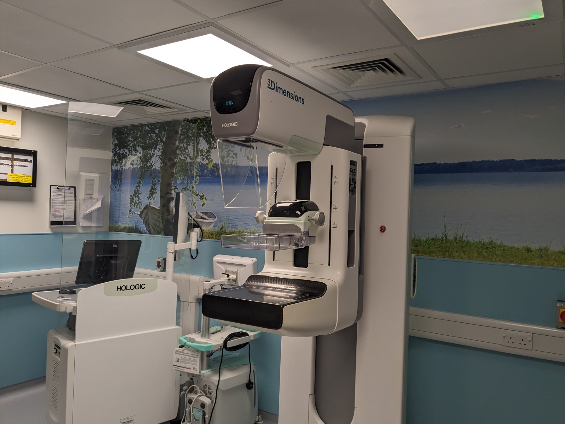 Photo of mammogram machine. An upright machine in smooth metal and plastic. There is a part that can be set to different heights, with a smaller part on that, also adjustable in height. This has a clear plastic part looking like a tray projecting out from it. The staff workstation is nearby behind a clear plastic screen.