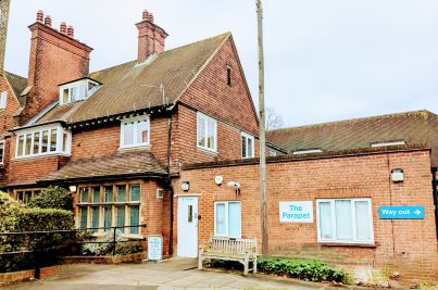 Photo of the parapet unit. A red brick house, perhaps 100 years old. There is a more modern small brick extension which has signage on the front and the entrance door. The external surface is paved and there's a wooden bench just outside the door.